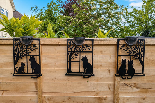 Decorative metal wall art of cats on a wooden fence with plants and trees in the background.